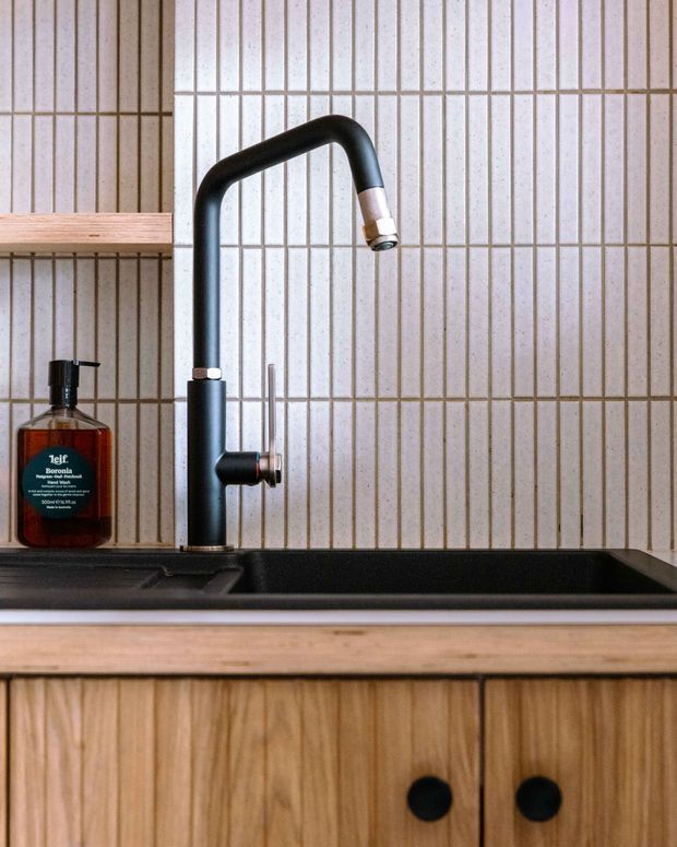 Close-up view of a compact kitchen sink area with a matte black gooseneck tap, deep black inset sink, timber benchtop, vertical timber cabinetry with round black knobs, and a cream tiled splashback with dark grout. A soap dispenser and simple timber shelf are also visible.