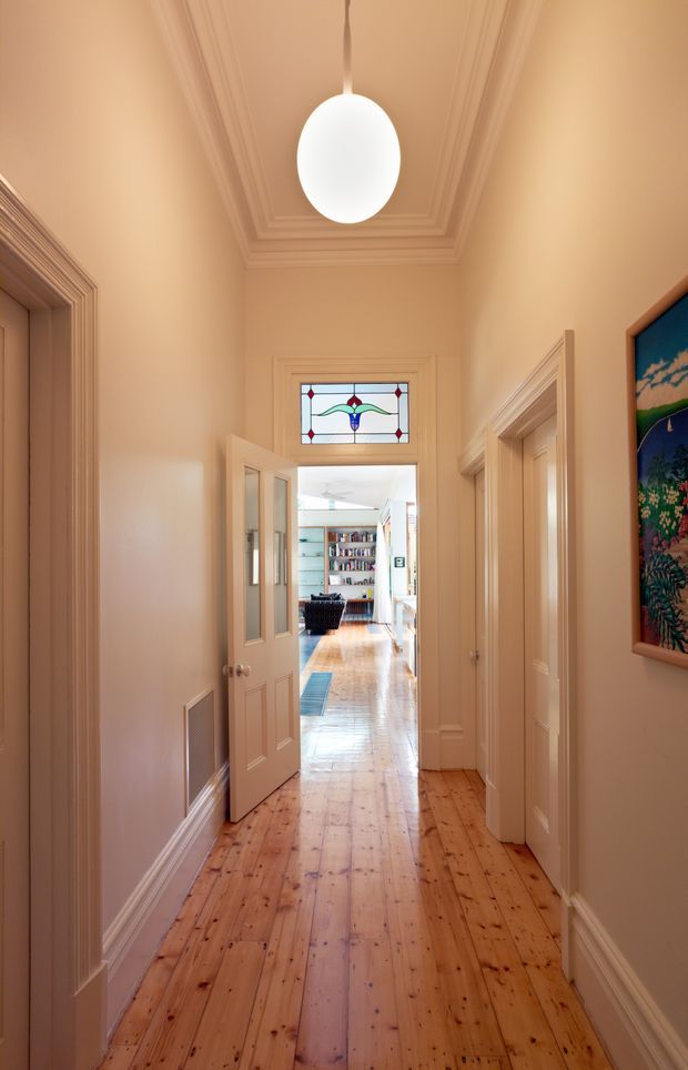 A narrow residential hallway with warm peach-beige walls, polished timber floorboards, ornate layered ceiling cornices, and detailed timber door architraves. A white oval pendant light hangs centrally, while a decorative stained-glass transom window sits above an open door leading into a bright living area with bookshelves and seating. Framed artwork is visible on the right wall.