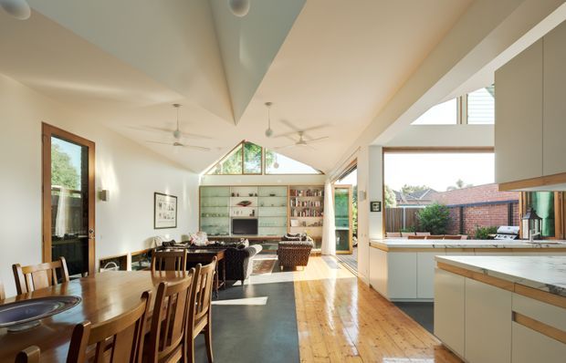 A bright, open-plan residential interior combining a dining area, living space and kitchen. The room has a high pitched ceiling with exposed geometric ceiling forms, ceiling fans, clerestory glazing and large windows. Warm timber flooring and timber-framed doors contrast with white walls and pale built-in cabinetry. A long dining table occupies the foreground, while a kitchen with a stone benchtop and outdoor-facing window is visible to the right. Built-in shelving and seating define the living area at the rear.