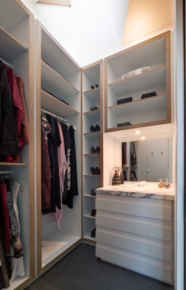 A compact custom walk-in wardrobe with floor-to-ceiling open shelving, hanging rails filled with clothing, dedicated shoe shelving, and a central built-in vanity. The vanity has a white marble-look benchtop, integrated drawers, illuminated mirror, and recessed downlights. Light timber edging frames the pale cabinetry, while dark carpet provides contrast. The space is brightly lit and efficiently organised.