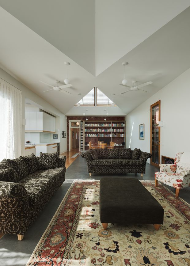 A bright, open-plan living room with a dramatic pitched, angular ceiling and clerestory glazing. The room features patterned dark sofas, a large traditional Persian-style rug, a black upholstered ottoman, a floral armchair, ceiling fans, and a full-height brick bookshelf wall. A compact white kitchen is visible to the left, with timber-framed doors and polished concrete flooring throughout.