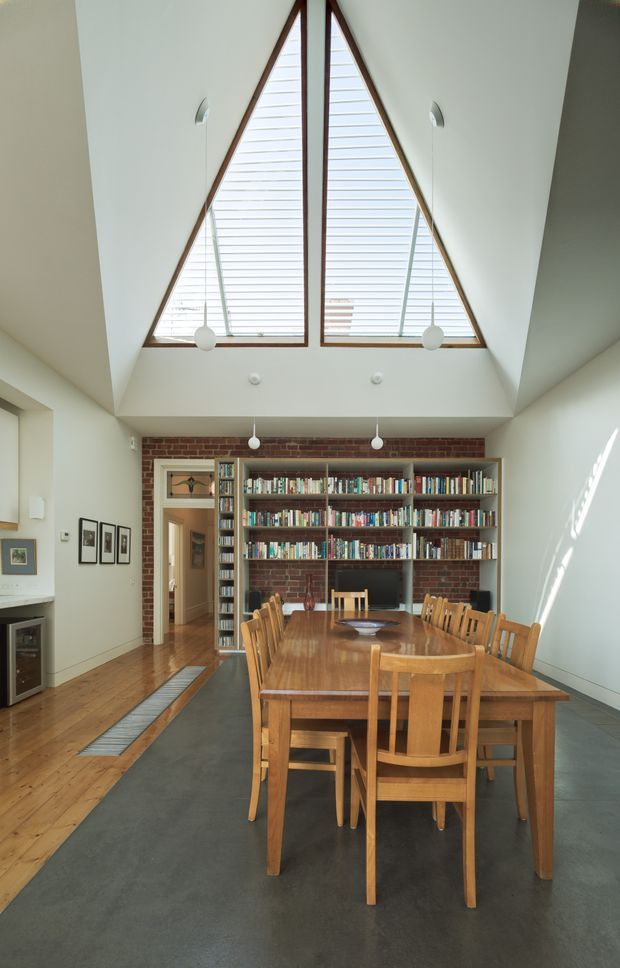 A bright, double-height dining and library space with a large timber dining table and matching chairs in the foreground. The room features a dramatic triangular clerestory window with timber framing, white angled walls and ceiling planes, exposed red brick shelving wall, extensive built-in bookcases, timber flooring at the perimeter, and a dark polished concrete floor through the dining area. Pendant lights hang above the table, with a hallway visible beyond the library.