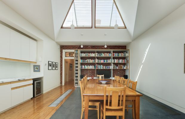 Bright open-plan dining area with a large timber dining table and matching chairs, set in front of a full-height brick feature wall with built-in bookshelves. A compact white kitchen with timber cabinetry and dark appliances occupies the left side. The room has polished timber flooring, a high pitched ceiling with triangular clerestory windows, and simple pendant lights. The restrained palette of white, natural timber, grey, and exposed red brick creates a warm, airy, library-like space.
