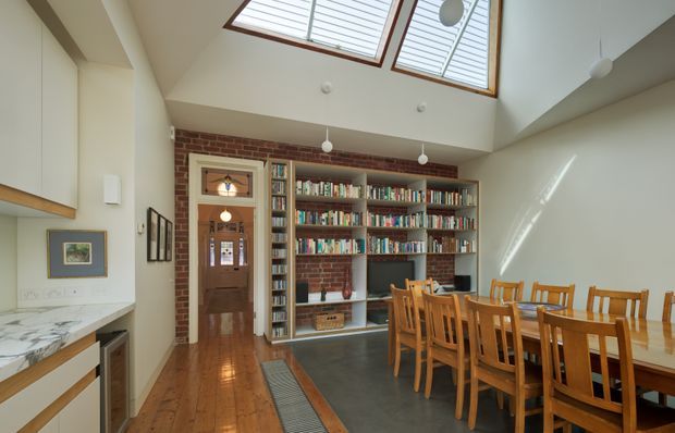 A bright, open-plan dining and living area with a long timber dining table and matching chairs in the foreground. Along the rear wall is an extensive built-in bookcase and media shelving set against exposed red brick, with a timber-framed doorway leading to a hallway. The room combines polished timber flooring with a dark tiled or concrete zone, pale walls, pendant lights, and large roof skylights that introduce abundant natural light. A partial kitchen or benchtop area is visible at left.