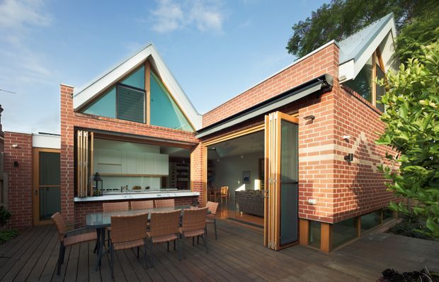 A compact brick-clad courtyard-style home with steep gabled roof forms, large triangular glazing, and wide openings connecting the interior to a timber deck. The outdoor dining area sits beside an open kitchen or living space, with timber-framed folding doors and a bright, contemporary architectural composition.