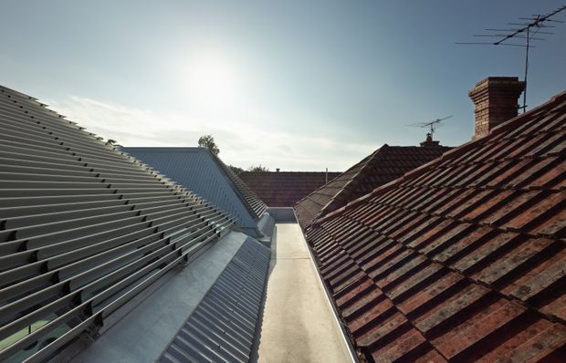 Exterior rooftop view looking along a narrow roof valley between a modern grey metal roof with pronounced horizontal ribbing and adjacent older reddish-brown tiled pitched roofs. Brick chimneys and rooftop antennas are visible against a bright sky.