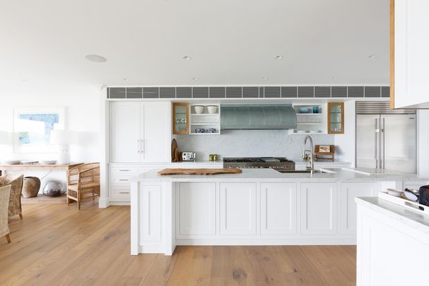 Bright, spacious open-plan kitchen with a large white paneled island, white cabinetry, pale stone benchtops, stainless-steel appliances, a grey tiled splashback, and timber flooring. A casual dining or living area is visible to the left, with abundant natural light and a clean coastal feel.