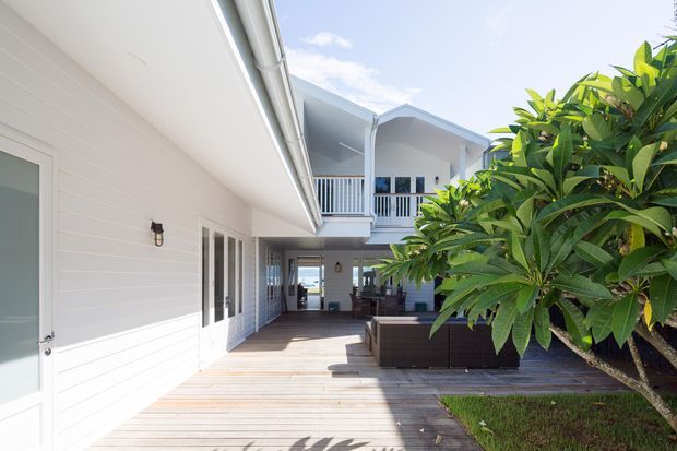 Bright exterior view of a white weatherboard-style multi-level residence arranged around a timber deck courtyard. The image shows covered outdoor living, an upper-level balcony with white balustrading, white-framed doors and windows, and tropical landscaping including a frangipani-like tree. Outdoor seating is visible beneath the covered area.