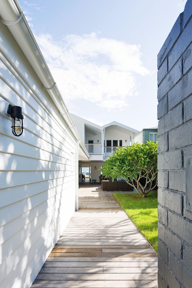 Bright exterior view along a timber deck walkway between a white weatherboard-clad building and a grey painted masonry wall, leading toward a raised residential structure with a covered balcony. The space includes white gutters, an exterior wall light, lush garden planting, and strong sunlight with soft shadows.