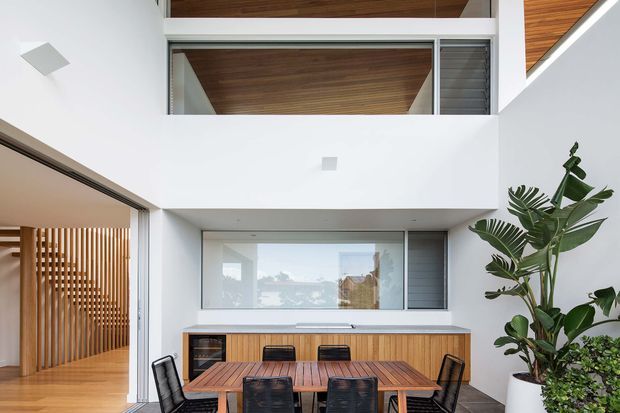 Bright contemporary dining area in a double-height courtyard-like interior, with a timber dining table and black chairs, white walls, extensive glazing, a vertical timber slat screen, and warm timber ceiling lining. Large indoor plants add greenery to the space.