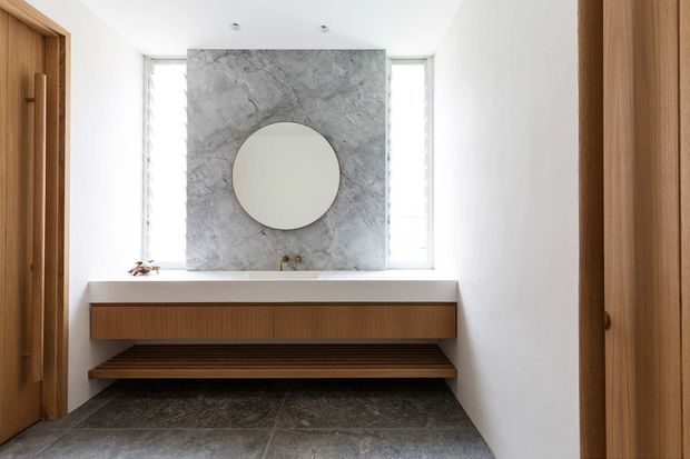 Minimalist entryway with white walls, a dark stone-tile floor, and a floating timber console with an open slatted shelf. A large grey veined stone feature panel with a circular mirror is framed by narrow windows, while timber doors and cabinetry add warmth.