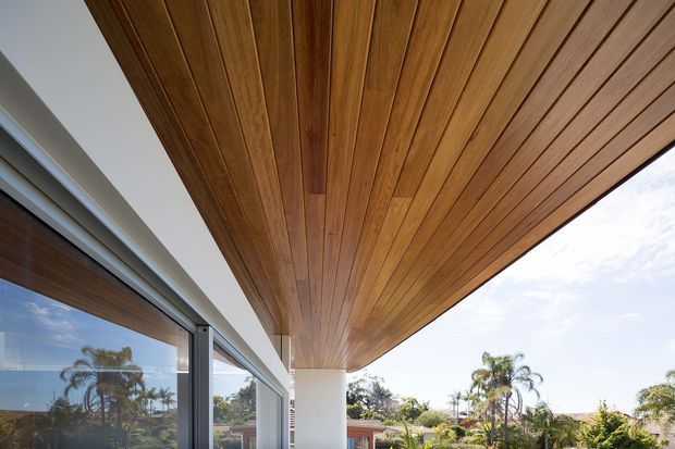A close-up exterior architectural view beneath a broad timber-lined soffit or roof overhang. The warm wood ceiling extends over a covered outdoor area, supported by a clean white structural column, with large dark-framed glazed sliding doors at left and landscaped palms visible beyond.