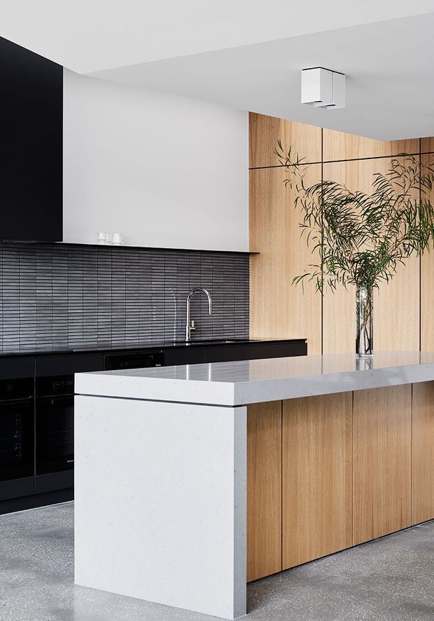 A contemporary minimalist kitchen featuring a large island with a pale stone benchtop and waterfall end, warm vertical timber cabinetry, glossy black lower cabinetry, and a dark horizontal tile splashback. A sculptural black-and-white pendant light hangs above, while a tall indoor tree adds organic contrast.