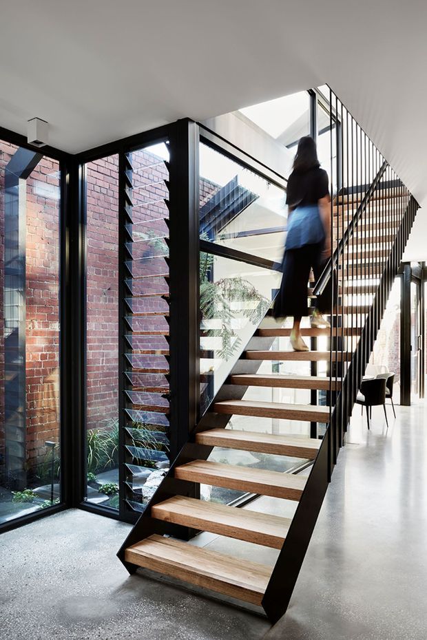 Interior architectural photograph dominated by an open-riser timber staircase with black metal stringers and slender black balustrades. The stair rises through a bright, double-height space beside full-height glazing and black-framed louvred windows. Exposed red brick walls and a small planted courtyard are visible through the glazing, with a dining area beyond. The space has an industrial-contemporary character, strong natural light, and a polished concrete floor.