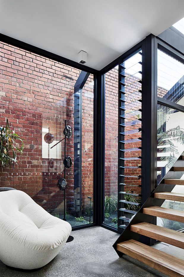 A contemporary interior corner at the base of an open-riser staircase, featuring exposed red brick walls, black-framed floor-to-ceiling glazing, dark metal stair structure with timber treads, and a sculptural cream lounge chair. A glazed shower-like display or water feature is visible against the brick wall, with louvred glazing and an adjoining courtyard-like view beyond.