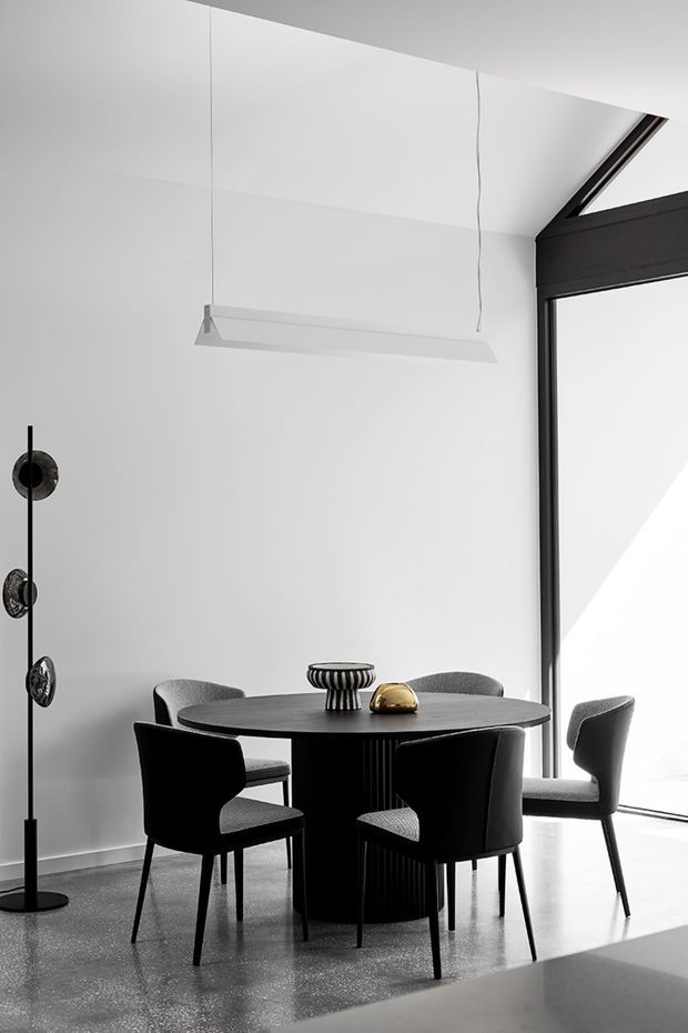 A refined contemporary dining area with a round black dining table and four upholstered dining chairs. The room has white walls, a polished grey concrete floor, a slim suspended linear pendant light, black-framed floor-to-ceiling glazing, and a sculptural black floor lamp. The restrained monochrome palette is accented by small metallic decorative objects on the table.