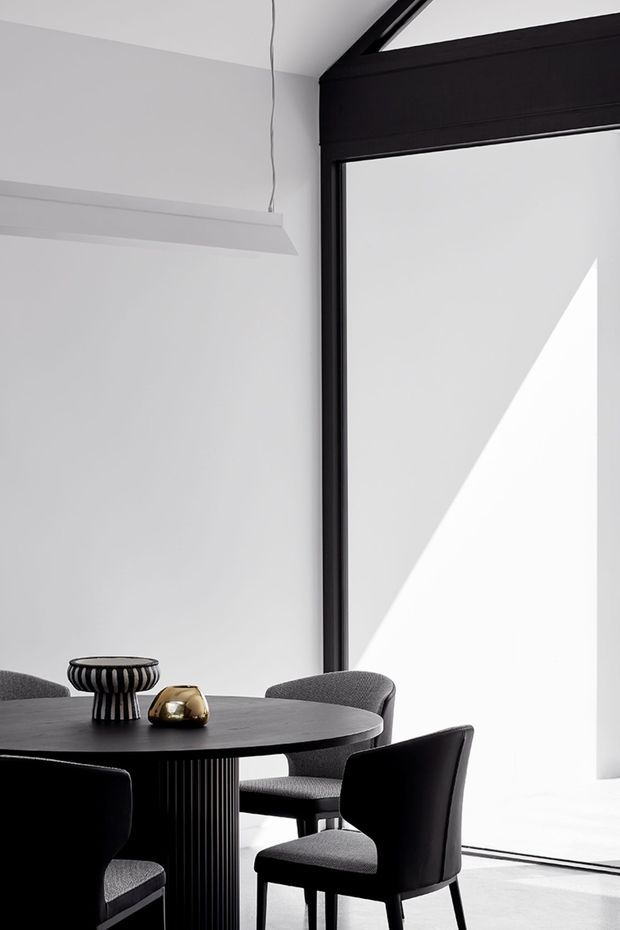 A minimalist dining area with a round dark dining table and sculptural pedestal base, surrounded by dark upholstered chairs. The room has white walls, a large black-framed glazed opening, a simple suspended linear pendant, and strong geometric sunlight creating a triangular shadow across the wall and floor. The palette is monochromatic with black, white, and charcoal-grey elements.