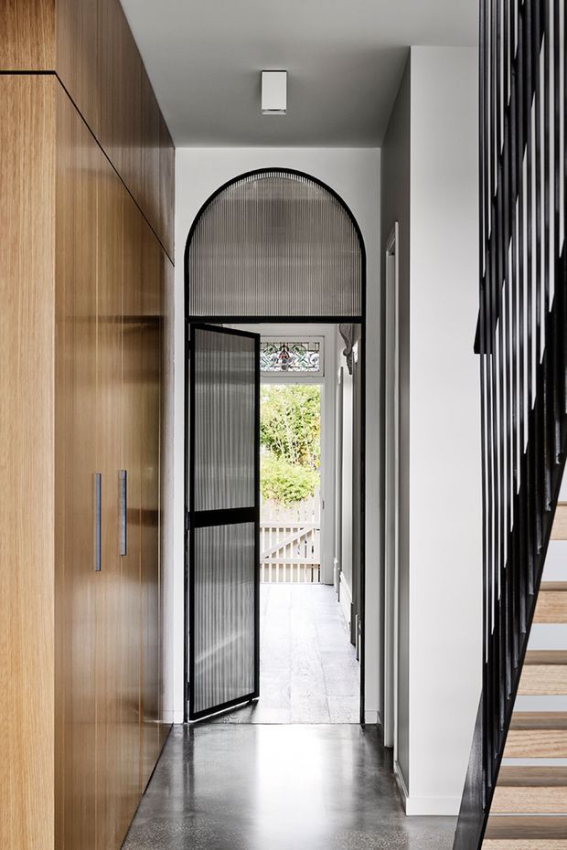 A narrow, refined residential hallway with full-height warm timber cabinetry on the left, a polished dark concrete floor, white walls and ceiling, and a black metal stair balustrade with timber stair treads on the right. At the end, a distinctive arched black-framed fluted-glass door opens toward a bright exterior porch and garden. The space combines clean contemporary lines with a heritage-style arched opening and decorative glazing.