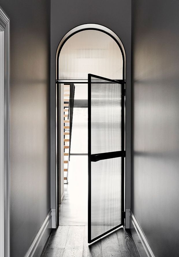 A narrow, refined hallway with dark grey walls, white decorative skirting and trim, and pale timber flooring. The focal point is a tall arched doorway with black metal framing and translucent ribbed glass panels, including a partially open pivot-style door. A bright corridor and timber stair elements are visible beyond. The image has a high-contrast, contemporary feel with traditional moulding details.