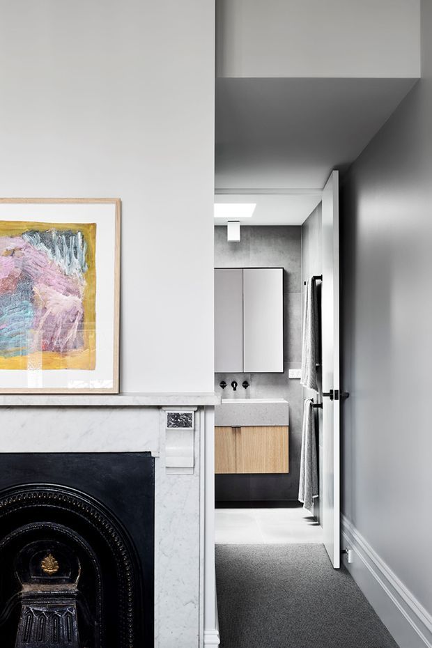 A refined residential hallway viewed beside a traditional fireplace, leading directly into a compact contemporary bathroom. The hallway has pale grey walls, white decorative skirting and trim, dark carpet, a marble fireplace surround, and framed abstract artwork. The bathroom features a grey concrete-look wall finish, a wall-mounted mirrored cabinet, a pale stone benchtop, timber vanity cabinetry, dark lower joinery, grey floor tiles, and black fixtures. The image combines heritage detailing in the corridor with restrained contemporary bathroom design.