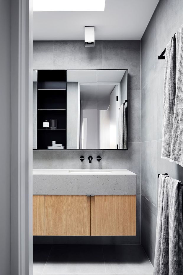 A compact, contemporary bathroom viewed through a doorway, featuring dark grey large-format wall tiles, a black floor, a floating natural wood vanity with a pale speckled stone benchtop, black wall-mounted tapware, and a mirrored cabinet with open black shelving. A skylight and ceiling vent provide overhead detail, while textured grey towels hang on the right wall.