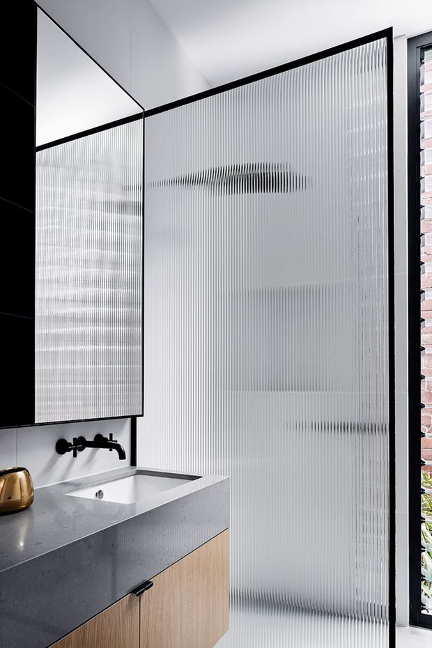 A compact contemporary bathroom vanity with a pale stone benchtop, integrated white basin, black wall-mounted tapware, and light timber cabinetry. A large ribbed translucent glass screen dominates the room, framed in black, with a full-height mirror and a narrow view of exposed brick and greenery beyond the window. The palette is monochromatic with warm timber and brass accents.