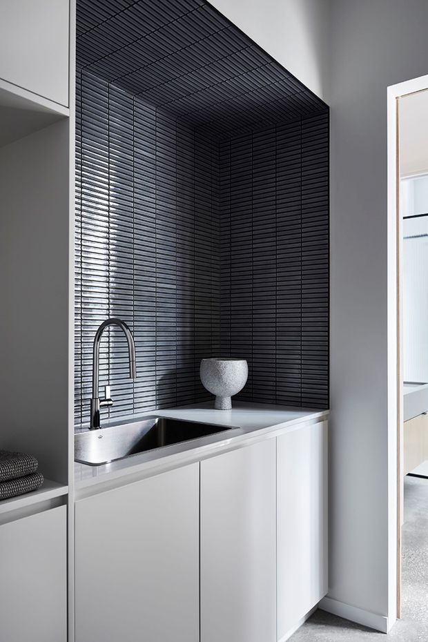 Compact contemporary utility space with a white built-in cabinet run, integrated stainless-steel sink and tall curved tap. The walls and ceiling are clad in a dramatic black narrow-ribbed tile or panel finish, contrasting with the white cabinetry and pale countertop. An open doorway reveals part of an adjacent room, while folded towels sit on open shelving at left.