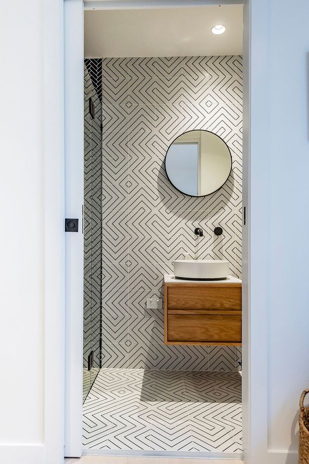 Compact modern bathroom viewed through an open doorway, featuring a bold geometric black-and-white patterned tile installation across the floor and walls, a frameless glass shower enclosure, a round wall mirror, wall-mounted black fixtures, and a floating warm timber vanity with a white vessel basin. The space is bright and graphic with clean contemporary detailing.