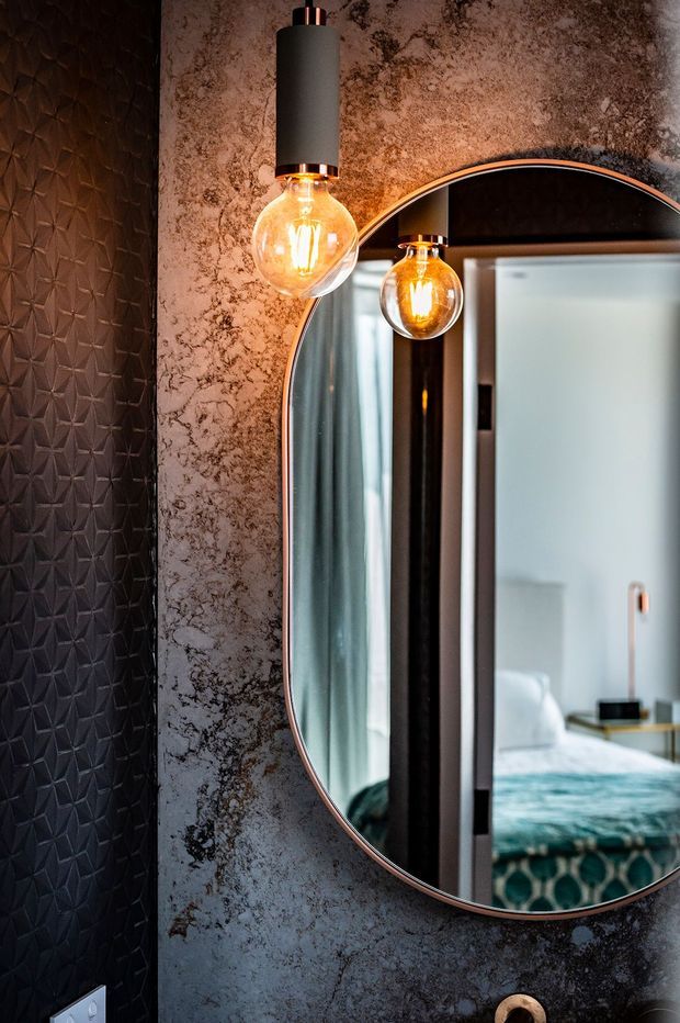 Close-up interior detail of a dark, moody bathroom or dressing area featuring a large rounded-rectangle mirror with a thin metallic frame, a textured black geometric wall surface, mottled dark stone-look wall finish, and a suspended cylindrical pendant light with an exposed warm Edison bulb. The mirror reflects a bedroom with a bed and soft blue-green bedding, indicating the bathroom or vanity area opens directly toward the bedroom.