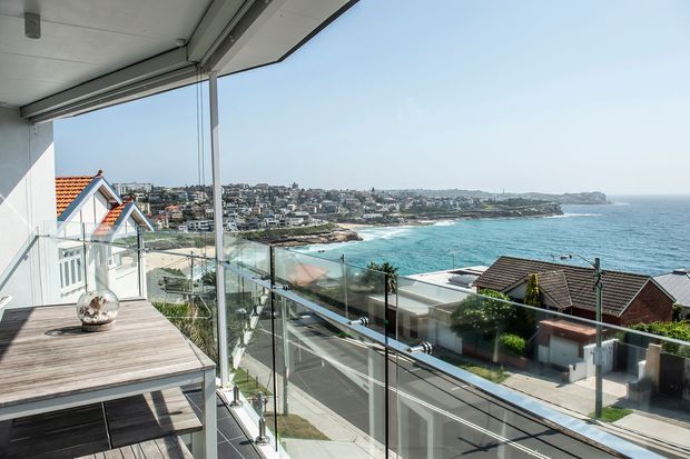 A bright coastal architectural view from a covered balcony or terrace, looking across a residential hillside neighbourhood toward a turquoise ocean and rocky shoreline. The foreground features a timber outdoor dining table, frameless glass balustrades, a glazed corner, and a contemporary roof overhang. The scene has strong expansive coastal and waterfront character with clear views toward the beach and headland.