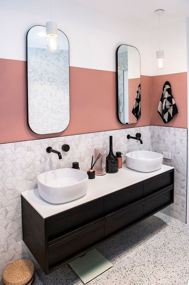 A contemporary bathroom featuring a double floating vanity with white vessel basins, matte black wall-mounted taps, black-framed arched mirrors, and pendant lighting. The walls combine a salmon-pink painted upper section with a white geometric mosaic tile splashback. Dark timber-toned drawers contrast with the white benchtop, while a terrazzo-style floor completes the bold, refined fit-out.
