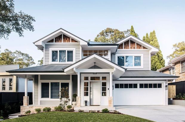 Front exterior view of a large two-storey suburban home with a symmetrical gabled form, white horizontal weatherboard cladding, dark metal roofing, prominent covered entry porch, shuttered windows, and an integrated double garage. The landscaped front yard and driveway are visible.