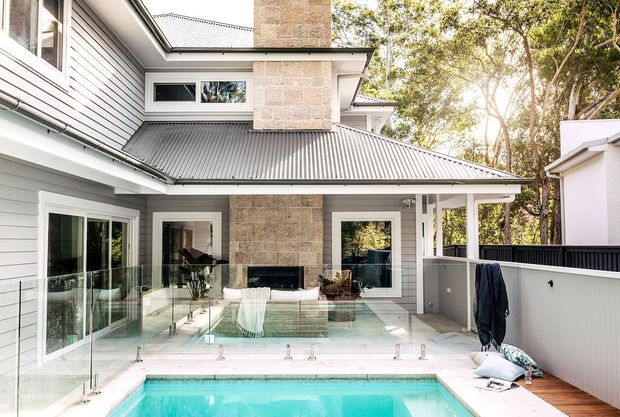 A bright residential courtyard viewed from beside an in-ground swimming pool. The house has light grey horizontal weatherboard cladding, white-framed glazing and doors, a dark corrugated metal roof, and a prominent beige stone-clad chimney with an outdoor fireplace. Glass pool fencing, timber decking, and mature trees surround the compact pool area.