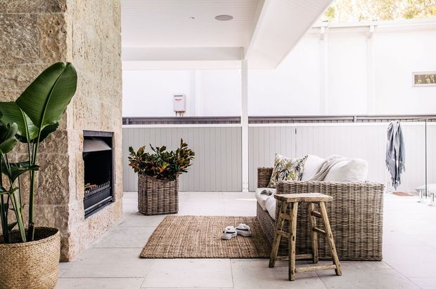 Bright covered outdoor living area with a pale tiled floor, white ceiling and walls, and a large rough-stone fireplace wall on the left. Wicker lounge furniture, woven rug, planter, tropical foliage, and a small side table create a relaxed entertaining space. The area appears enclosed or sheltered by a lightweight white structure with high-level glazing or openings.