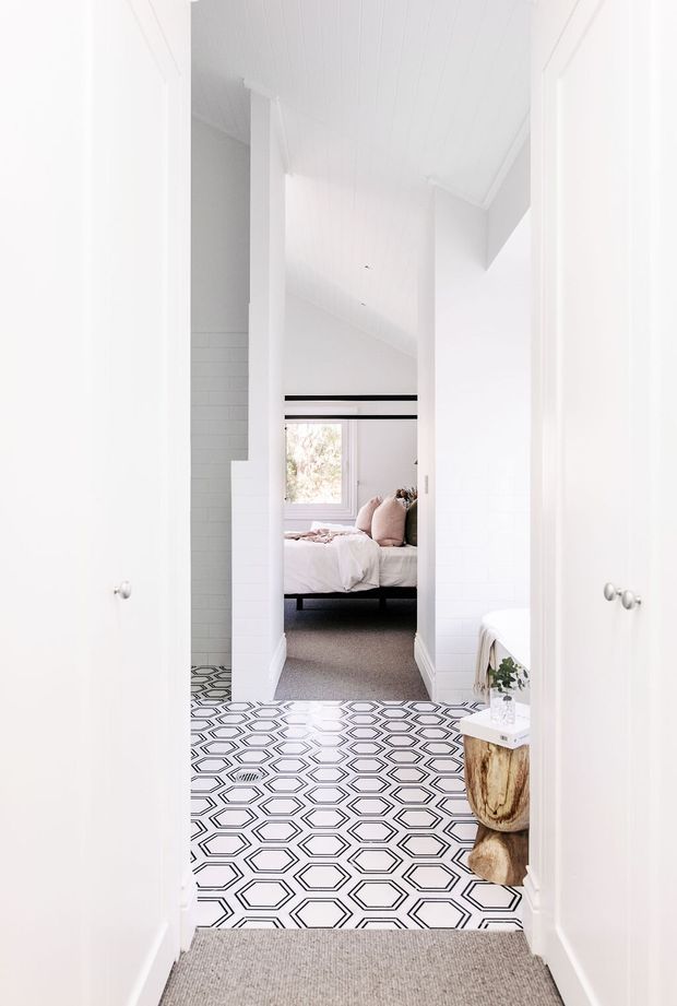 Bright white hallway or landing viewed through open doors, featuring a bold black-and-white geometric tiled floor, white paneled walls and cabinetry, and a high white ceiling. A bedroom with a bed and soft neutral bedding is visible at the end of the corridor, while part of a white bathtub or bathroom fixture appears on the right. The space has a clean contemporary feel with contrasting patterned flooring and natural wood accents.