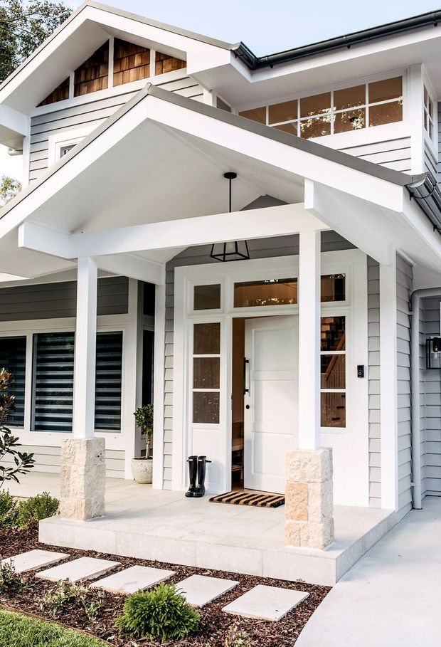 A bright exterior view of a two-storey suburban home entry. The facade combines white-painted horizontal weatherboards with darker grey weatherboard sections, a covered gabled porch supported by square white posts, pale stone-clad column bases, large white-framed windows, and a glazed front door. A simple landscaped path with square stepping stones leads to the porch.