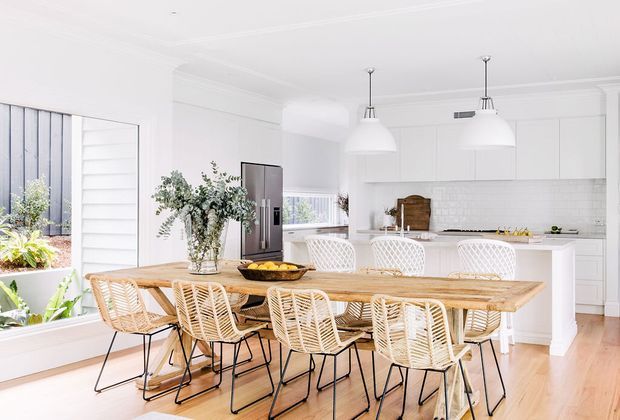 Bright, spacious open-plan kitchen and dining area with white shaker-style cabinetry, white subway-tile splashback, a large white island, stainless-steel refrigerator, and pendant lights. A long natural timber dining table is surrounded by woven rattan chairs, with large windows and indoor plants bringing in abundant daylight. The room has a clean, relaxed coastal feel.