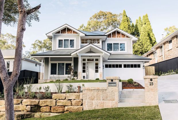Street-facing view of a large two-storey suburban residence with a symmetrical gabled facade, white horizontal weatherboard cladding, dark grey pitched roofs, white-framed windows with shutters, and a covered entry porch. The property includes an integrated double garage, front retaining walls in pale stone, landscaped planting beds, steps, and a driveway. Mature trees and neighbouring homes frame the exterior.