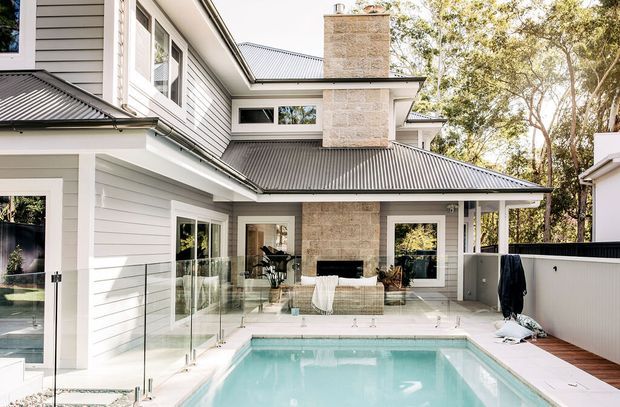 A bright rear exterior view of a multi-level home centred around a compact swimming pool and terrace. The house has pale horizontal weatherboard cladding, light masonry feature walls and chimney, dark corrugated metal roofing, extensive glazing, and a glass pool balustrade. Timber decking, outdoor furniture, and mature trees surround the pool courtyard.