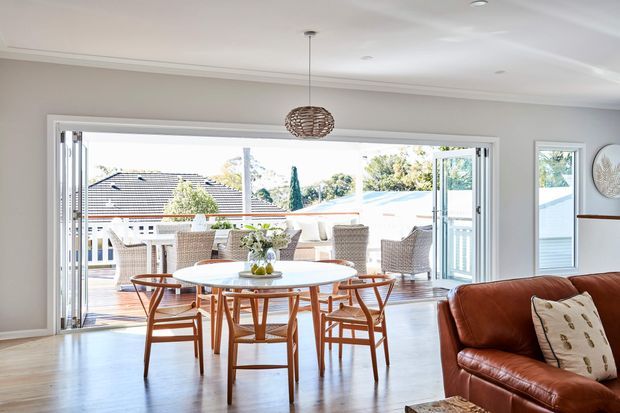 Bright open-plan dining and living area with a round timber dining table and chairs positioned beside expansive bi-fold glazing. The room has pale neutral walls, a polished timber floor, a woven pendant light, and a brown leather sofa. The glazing opens onto a furnished outdoor deck or terrace, creating a strong indoor-outdoor connection.