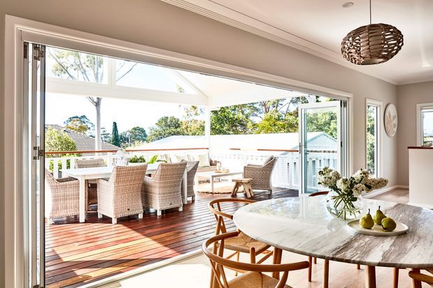 Bright open-plan dining area with a large round marble dining table and woven wooden chairs, opening through expansive sliding glass doors onto a timber deck with an outdoor dining setting. The room has warm neutral walls, white architectural trim, abundant natural light, and a woven pendant light. A leafy suburban exterior is visible beyond the deck.