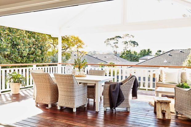 Bright covered outdoor entertaining area with a timber deck, white-painted posts and balustrades, and a dining table surrounded by woven wicker chairs. A cushioned outdoor seating area and potted plants sit to the side, with neighbouring pitched-roof homes visible beyond the railing.