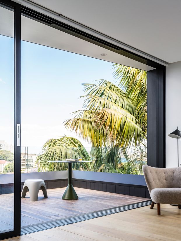 A bright contemporary interior opening onto a balcony or terrace through fully retractable black-framed glazing. The room has pale timber flooring, a dark built-in window base and trim, a sculptural green pedestal side table, a small white stool, and a textured upholstered armchair. Tropical palm foliage and a distant urban setting are visible outside.