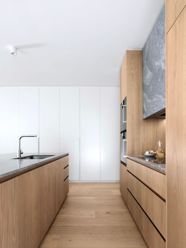 A bright, minimalist contemporary kitchen viewed down a central aisle. The space features full-height white cabinetry at the rear, a large wood-panelled island with an integrated sink and slim metal tap, and matching timber cabinetry along the right wall. The right-side joinery incorporates built-in ovens, a stone benchtop, and a dramatic grey textured rangehood or splashback feature. Light timber flooring and clean white surfaces create an airy, refined appearance.