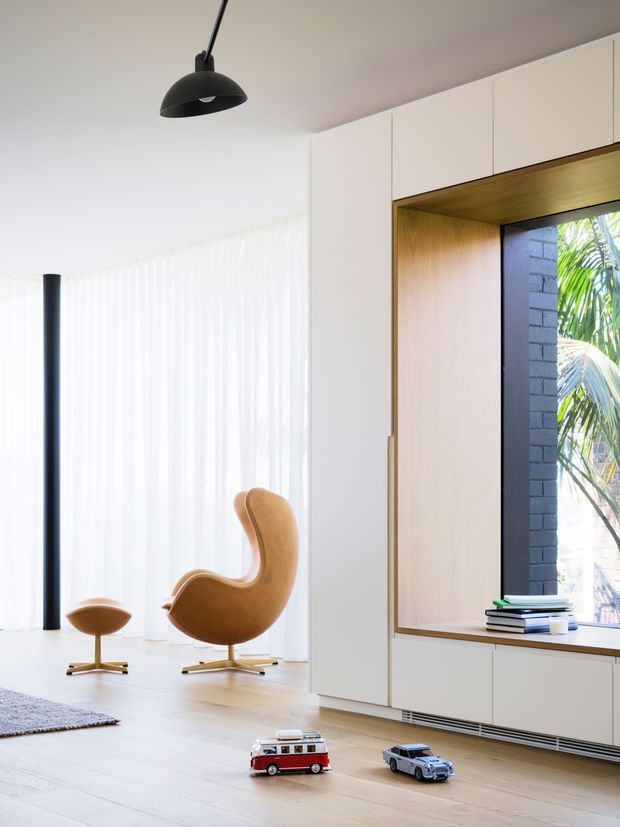 Bright, minimalist living room with full-height sheer curtains, pale timber flooring, white built-in cabinetry, and a timber-lined recessed window seat or display niche. A sculptural tan lounge chair, matching low chair, black pendant light, and slim black stove or architectural column provide contrast. Books and toy vehicles sit in the foreground, while greenery is visible outside the window.