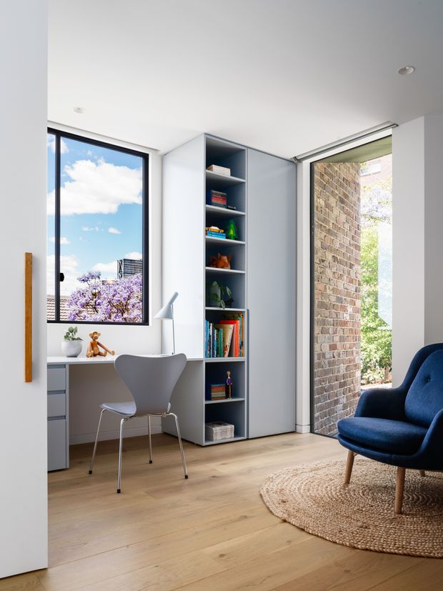 Bright, compact contemporary interior combining a built-in home office with a small reading or lounge area. A white desk with drawers sits beneath a large black-framed window, paired with a simple grey chair and desk lamp. Floor-to-ceiling grey built-in shelving provides open book storage and closed cabinetry. A deep blue upholstered armchair sits on a round natural-fibre rug beside a large glazed opening, with exposed brick visible outside. Pale timber flooring, white walls and abundant daylight create an airy, refined space.