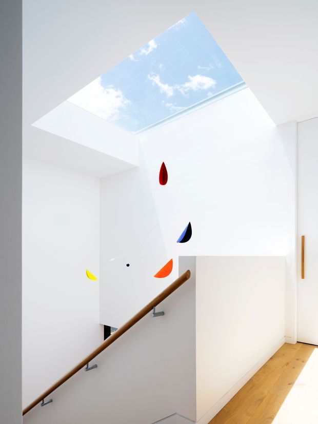 Bright minimalist stairway with white walls and a large rectangular skylight showing blue sky. A simple timber handrail runs along the stair, with small colourful abstract wall accents and light timber flooring on the landing.