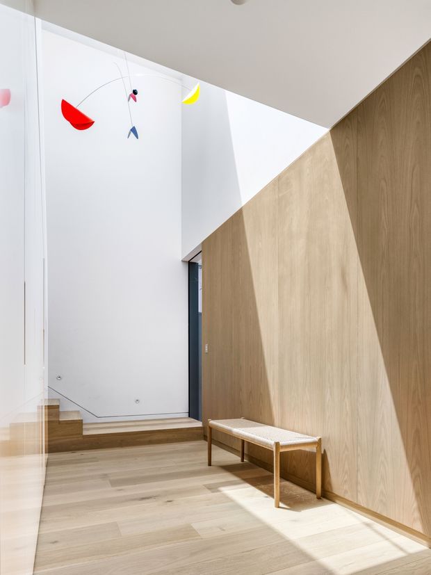 A bright, minimalist interior entry hall with a short set of timber stairs, pale timber flooring, and a full-height natural timber wall with geometric angled sections. A simple woven-seat timber bench sits along the wall, while a colourful suspended mobile hangs near the tall white wall. Strong sunlight creates angular shadows across the floor and wall.