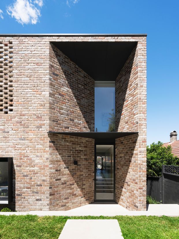 Front exterior view of a contemporary brick house with a tall recessed entry, deep angular brick reveals, a dark flat canopy, and narrow full-height glazing. The facade uses varied warm pale and reddish brickwork with strong geometric shadow lines, set behind a simple lawn and concrete path under a clear blue sky.