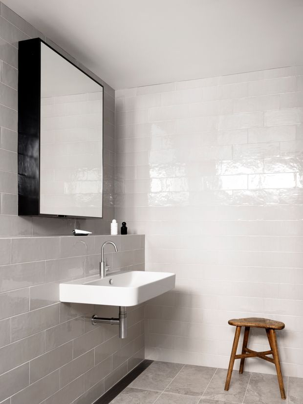 A minimalist bathroom with a wall-mounted white rectangular basin, exposed chrome plumbing, a simple curved faucet, and a large black-framed mirrored cabinet. The walls and raised vanity ledge are clad in glossy white rectangular tiles, while the floor uses large matte grey tiles. A small wooden stool adds warmth against the otherwise monochrome palette.